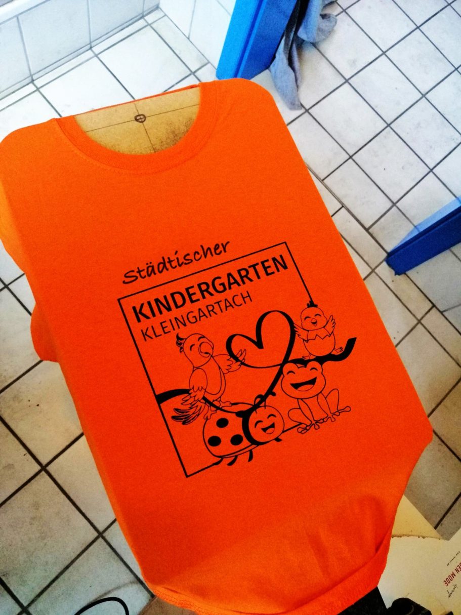 Bringen Sie Farbe in den Kindergartenalltag! Auf diesem Bild sehen Sie ein fröhliches orangefarbenes Kinder-T-Shirt, das mit einem liebevollen Design für unseren Kindergarten bedruckt wurde. Mit unserem hochwertigen Siebdruckverfahren stellen wir sicher, dass jedes Shirt nicht nur toll aussieht, sondern auch langlebig und bequem ist. Ideal für Ausflüge, Veranstaltungen oder einfach für den Alltag – unsere T-Shirts fördern den Gemeinschaftsgeist und machen das Spielen noch schöner!